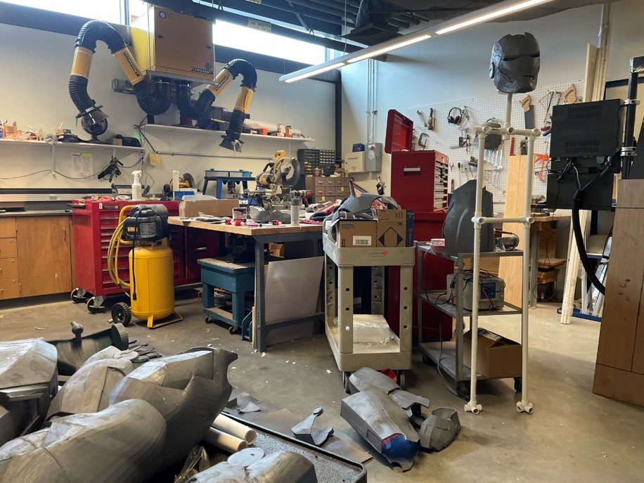 Workshop with printed gray armor pieces on the floor and a PVC pipe frame holding the helmet